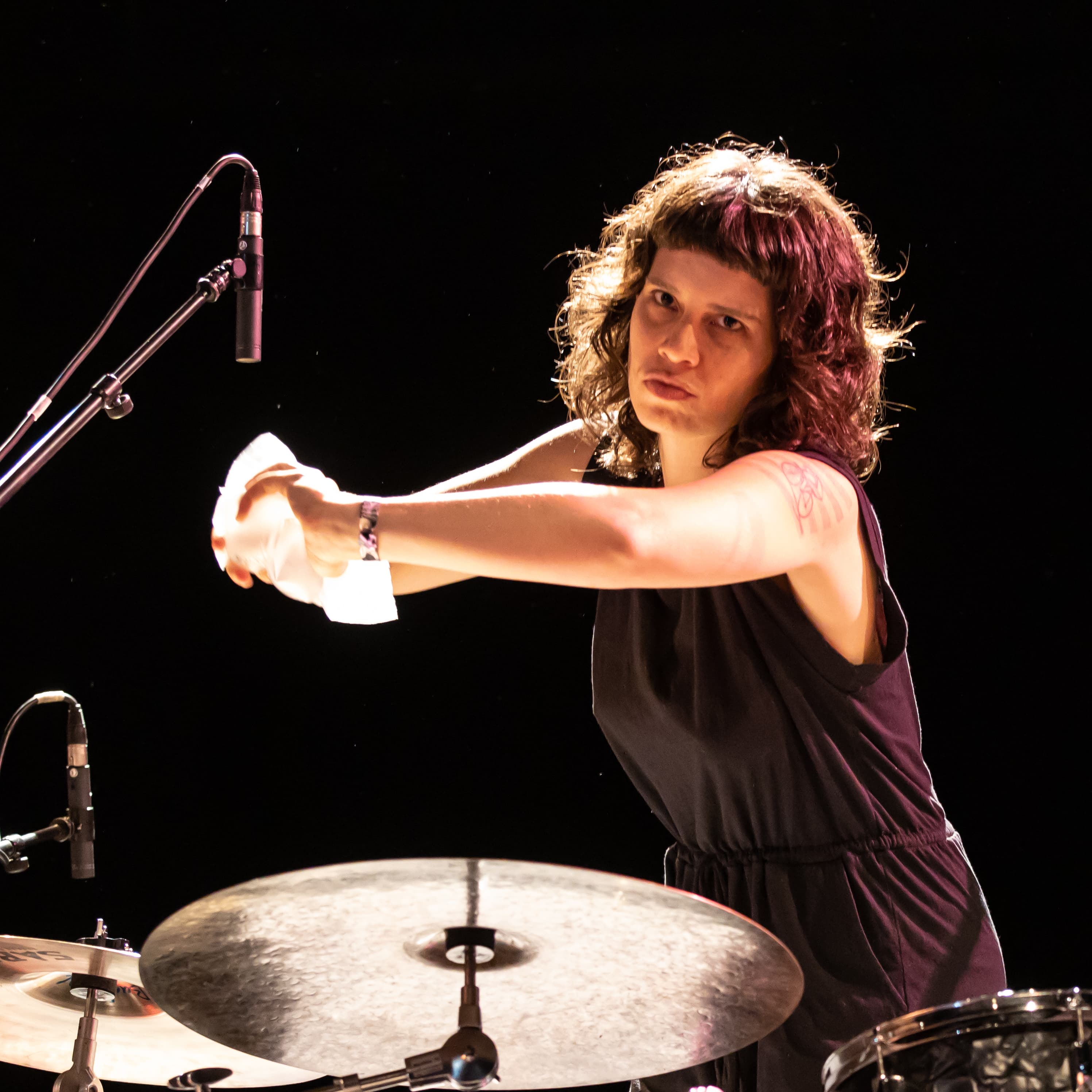 A person with curly hair and a tattoo on their arm is playing drums under stage lighting.