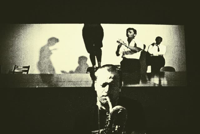 A black-and-white scene shows a musician playing a saxophone in the foreground, with a projected image of people and their shadows in the background.