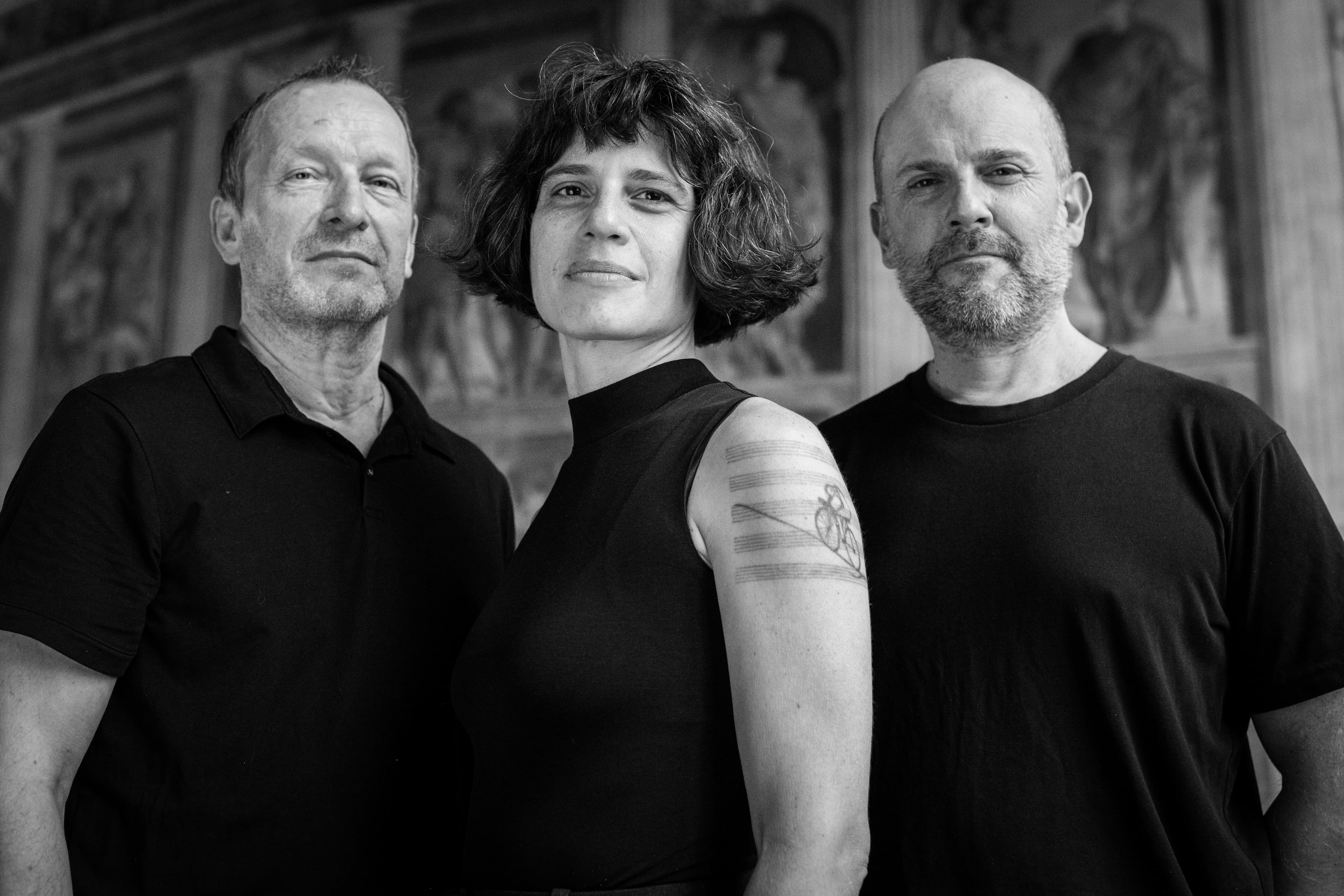 Three people stand close together, looking confidently at the camera in a black and white setting.