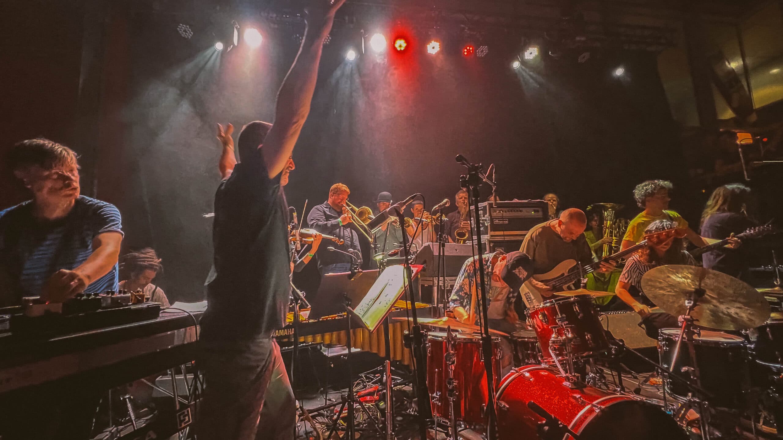 A dynamic live band performance featuring multiple musicians playing instruments like drums, guitar, and brass, under vibrant stage lighting.